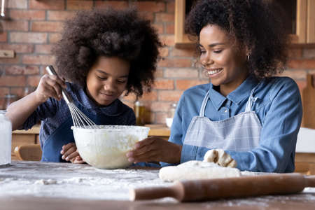 Loving smiling young African American mother teach small teen daughter cooking making dough together...