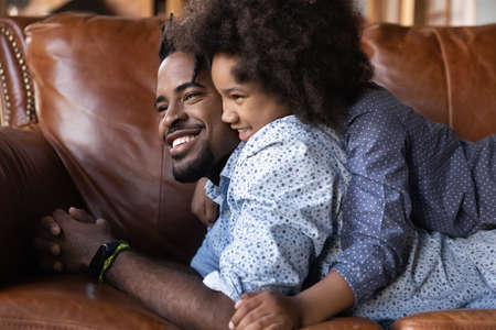Cute happy little girl child lying on young African American father relax play together at home. Smiling ethnic dad and small daughter rest on sofa at home, enjoy family weekend. Fatherhood concept.の写真素材
