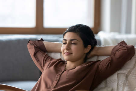 Calm happy millennial Indian woman relax in chair take nap or sleep relieve negative emotions. Peaceful young mixed race female rest at home breathe fresh condition ventilated air. Peace concept.の写真素材