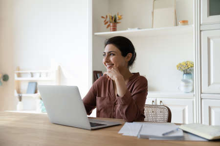 Happy millennial Indian woman sit at desk at home office work on laptop look in distance dreaming or...