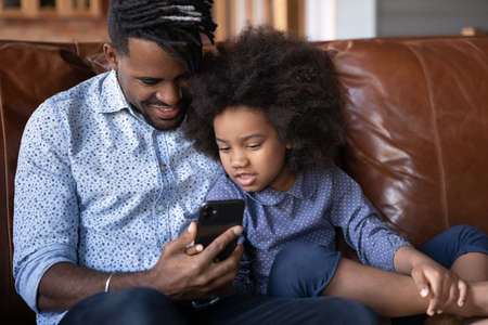 Loving happy young African American dad and little teen daughter rest on sofa use modern smartphone...