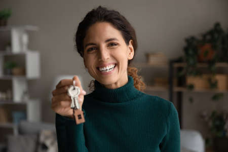 Head shot portrait smiling woman showing keys at camera, purchasing new house, moving into first new apartment, happy young female tenant renter rejoicing relocation, mortgage or rent conceptの写真素材