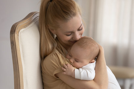 Happy young Caucasian mother hug caress little baby infant son or daughter sleeping in hands. Smiling mom sit in chair rest cuddle lull small newborn kid child relax in arms. Motherhood concept.の写真素材