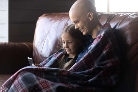 Happy ill mom and daughter kid relaxing in plaid on sofa, using mobile phone, making video call, watching virtual content, shopping online. Mom with cancer and child enjoying family time togetherの写真素材