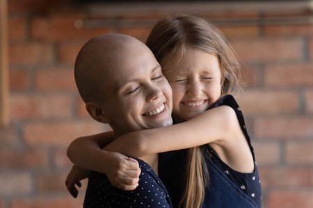 Happy daughter girl hugging mom with cancer, holding tight, smiling with closed eyes. Little kid giving love, motivation to ill hairless young mother during chemotherapy. Family support conceptの写真素材
