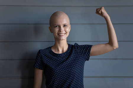 Happy strong cancer survivor beating disease, winning fight for life. Young woman with shaved head looking at camera, smiling, making power strength hand gesture. Head shot portraitの写真素材