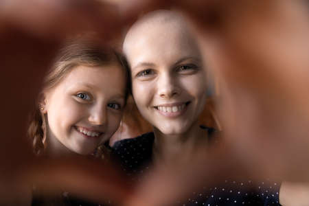 Happy young mom with cancer and cute little daughter kid having fun, smiling at camera through heart shaped hand frame. Optimistic patient enjoying leisure time with child. Close up portraitの写真素材