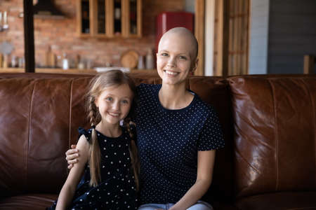 Happy mom with hairless head hugging cute little daughter girl at home, enjoying leisure time with kid on sofa, smiling at camera. Woman going through cancer with support of family, Headshot portraitの写真素材