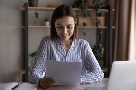 Happy millennial Caucasian woman sit at desk work on computer read good news in paperwork letter....