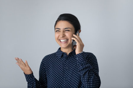 Overjoyed millennial Indian woman isolated on grey studio background laugh talking speaking on cellphone. Smiling young biracial female have fun enjoy pleasant smartphone communication online.の写真素材