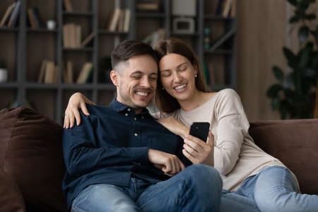 Happy 30s couple sit on sofa use smartphone enjoy new mobile app, carefree family spend time on...