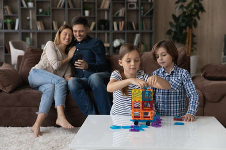 Kids sit at table play together plastic magnetic constructor while young parents use smart phone...