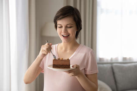Excited young expecting mom enjoying chocolate dessert. Smiling hungry pregnant woman eating slice of cake, standing in living room at home, using fork. Nutrition in pregnancy, sweet food conceptの写真素材
