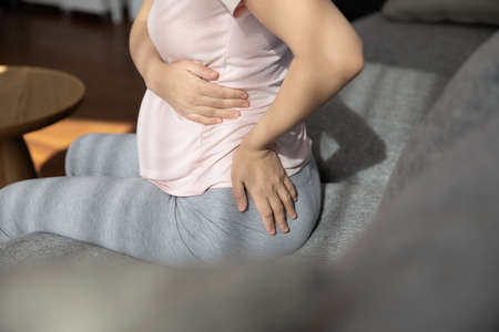 Late pregnant woman suffering from backache, sitting on couch, holding belly, touching back. Expectant mother tired of overweight, having health problems, feeling lumbago muscles pain. Cropped shotの写真素材