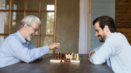 Smiling old grey-haired 60s father have fun play chess with young grownup son relax together on...