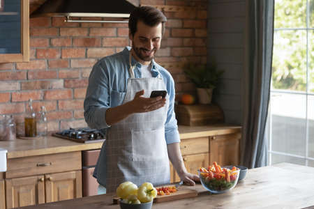 Smiling millennial Caucasian man in apron cook tasty salad for lunch check recipe on smartphone online. Happy young male prepare healthy tasty food for dinner, use cellphone texting on web.の写真素材