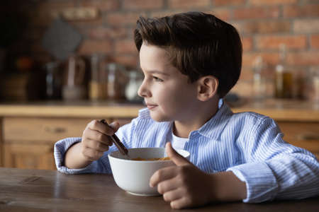 Happy small Caucasian boy sit at desk at home eat healthy cereals with organic milk for breakfast. Healthy little child kid enjoy tasty natural bio diet food in kitchen. Children healthcare concept.の写真素材