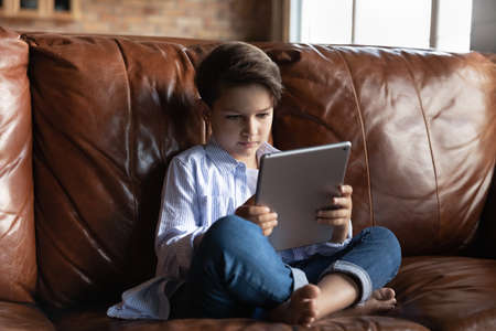 Serious small teen Caucasian boy kid sit on couch at home hold use tablet gadget study online. Smart...