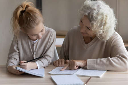 Loving mature Caucasian grandmother help little teen granddaughter with school homework assignment. Caring old grandparent and small grandchild study together prepare task. Education concept.の写真素材