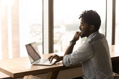 Smiling millennial African freelancer man working on laptop looks at device screen watch webinar...