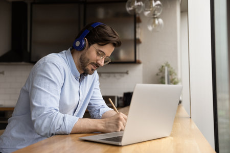 Young focused man student sit at table at home wear modern wireless headphones, listen audio course...