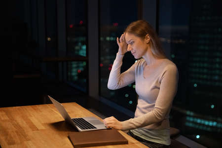 Serious workaholic tired bemused businesswoman work until late in office, sit at desk using laptop solve issues, feels puzzled overloaded by over hours job at workplace, night city view through windowの写真素材