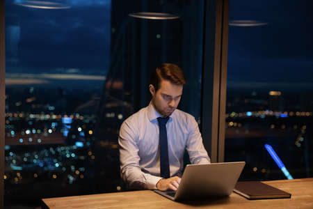 Confident ambitious corporate male office employee sit at workplace work until late use laptop, lead...