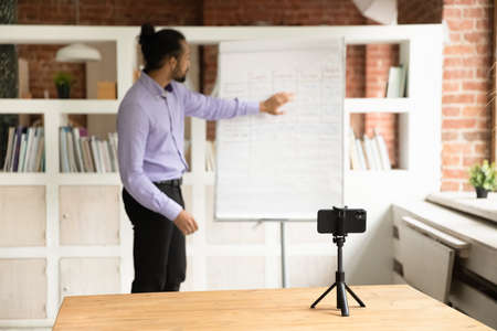African young male teacher blogger record webinar educational class use cell phone on tripod, explain written information pointing on flip chart. Improve language skills, tutoring, education conceptの写真素材