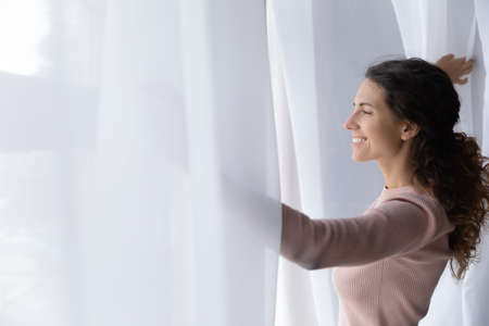 Young Latina woman open curtains at home or hotel looks out window thinking or dreaming, admire...