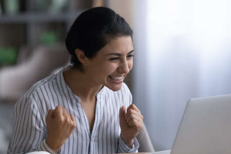 Overjoyed young Indian woman feel euphoric with online lottery win victory on laptop. Smiling happy...