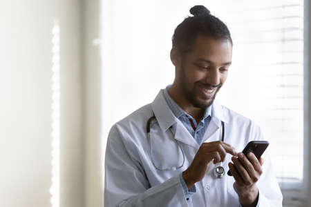 Smiling millennial generation african american biracial gp doctor physician using cellphone, reading message email with pleasant news, communicating online with patient or web surfing in clinic.の写真素材