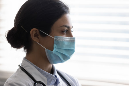 Close up head shot side view thoughtful young indian gp doctor therapist wearing facial protective mask looking out on window in distance, consider illness treatment, virus prevention measures.の写真素材