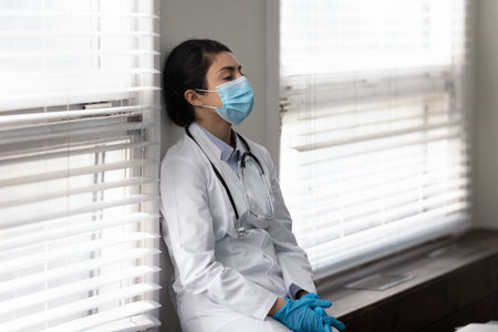 Stressed unhappy young indian ethnicity doctor in protective facemask and disposable gloves sitting on windowsill in clinic office room, feeling exhausted after hard workday in infectious department.の写真素材