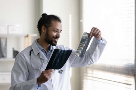 Happy young african american multiracial male surgeon traumatologist holding x-ray images, examining analyzing fracture in clinic, considering problem solution, making diagnosis, medicare concept.の写真素材