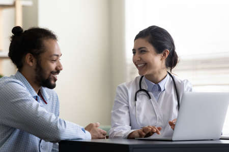 Happy millennial african american patient discussing computer presentation of medical insurance program with smiling skilled indian female general practitioner doctor physician at meeting in clinic.の写真素材