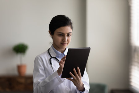 Concentrated young indian female doctor general practitioner therapist using applications on digital tablet, consulting patient online, prescribing treatment distantly, medicine and modern tech.の写真素材