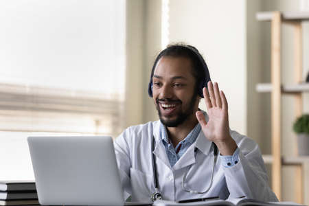 Happy friendly young african american biracial doctor in headphones waving hand, starting distant...