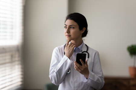 Thoughtful young indian female general practitioner doctor holding cellphone in hands looking in distance, considering problem solution or illnesses treatment consulting patient distantly online.の写真素材