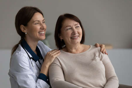 Kind affectionate young female doctor therapist psychologist cuddling shoulders of elderly senior smiling woman, looking together in distance, doing mental health exercises or dreaming at clinic visitの写真素材
