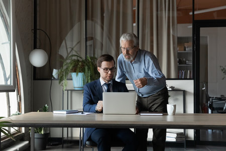 Senior businessman wearing glasses mentor instructing training new employee, using laptop, helping with corporate software, two colleagues working on online project together, discussing strategyの写真素材