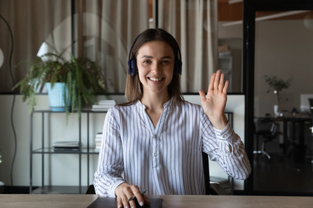 Head shot portrait smiling businesswoman in headphones waving hand and looking at camera, friendly employee worker involved in internet meeting, video call, happy mentor teacher leading online lessonの写真素材