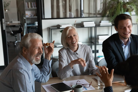 Excited mature businesspeople with colleagues eating pizza during break in office together, happy senior coworkers laughing at funny joke, talking chatting having fun, sharing corporate lunchの写真素材