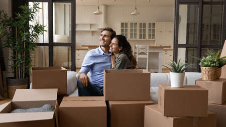 Dreamy young happy bonding family couple cuddling sitting on sofa near big carton boxes, visualizing future, planning decoration in renovated stylish living room, resting together in own home.の写真素材