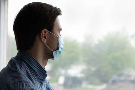 Thoughtful depressed millennial man looking out of window with hope, thinking over business lockdown...