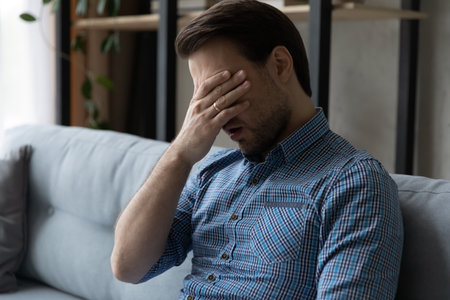 Tired bored millennial guy feeling exhausted, suffering from lack of sleep, laziness, low energy level, hangover. Man touching head, eye, feeling headache. Frustration, exhaustion, fatigue conceptの写真素材