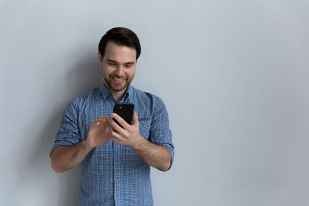 Happy millennial man using online app on smartphone, reading text message on mobile phone screen,...