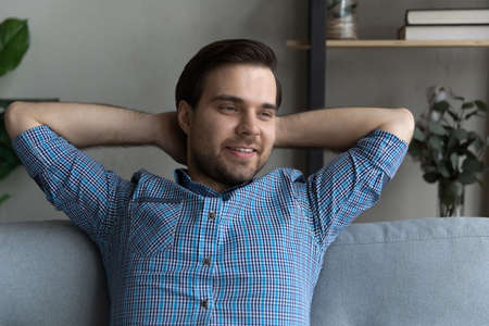 Thoughtful smiling guy relaxing on couch, looking away and smiling, thinking over achievement, planning good events. Millennial man resting on sofa at home, enjoying break, breathing fresh airの写真素材