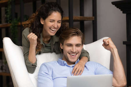 Happy young bonding family couple looking at laptop screen, celebrating getting email with good news. Emotional laughing spouses making yes gesture, reading amazing online lottery win notification.の写真素材