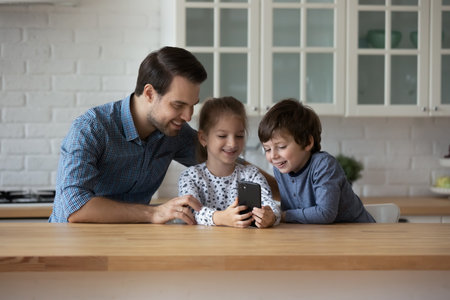 Happy dad and sibling children watching virtual content, chatting online on smartphone, shopping on...
