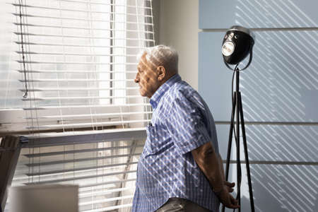 Thoughtful sad senior 80s man looking out of window, thinking over memory loss, dementia, Alzheimer...